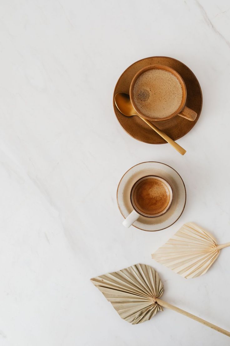 a cup of coffee next to two bamboo fans on a white counter top with gold colored spoons