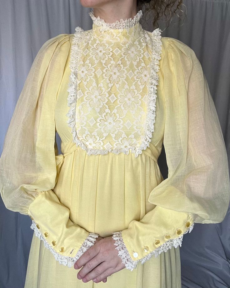 Details: maxi length; soft yellow; acetate lining with overlay; white lace trim toward bottom of skirt; around sleeve cuffs and at bodice; puffy, billowy, semi-sheer long sleeves with oversized button up cuff; lace bib detail; mock neck; attached front tie belt and zip up closure in the back.  Damage: minor separation under armpit seam on front lefthand sleeve; small stain on front layer toward hemline under lace trim; small spots on back hemline layer (well hidden in pleats); tears in acetate l Cream Long Sleeve Dress With Lace Collar, Flowy Lace Maxi Dress With Long Sleeves, Flowy Long Sleeve Lace Maxi Dress, Flowy Lace Long Sleeve Maxi Dress, Vintage Long Sleeve Dresses With Lace Patchwork, Long Sleeve Lace Maxi Dress With Lace Trim, Yellow Long Sleeve Maxi Dress With Ruffles, Yellow Long Sleeve Ruffled Maxi Dress, Spring Maxi Dress With Ruffles And Lantern Sleeves