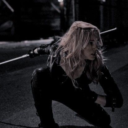 a woman kneeling down on the ground holding two swords in one hand and looking at something behind her