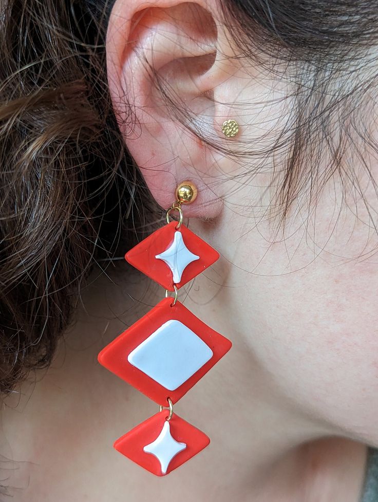 Step back in time with these retro red-orange and white polymer clay dangle earrings, perfect for adding a pop of color and vintage style to your outfit. Handmade with care, each earring is one-of-a-kind and features a unique retro design with a nod to the 60s and 70s. These statement earrings are made from high-quality polymer clay, which makes them lightweight and comfortable to wear all day long. The dangle style adds movement and dimension to the earrings, making them perfect for adding a touch of fun to any outfit. These earrings are perfect for those who love unique and quirky jewelry, and they make an excellent gift for the retro lover in your life. These earrings are sure to be a hit with anyone searching for statement jewelry that stands out from the crowd. Order your pair today a Star Polymer Clay, Mod Culture, Idea Lab, 60s Earrings, Cat Clay, 70s Earrings, Earring Shapes, 60s Jewelry, Clay Inspo