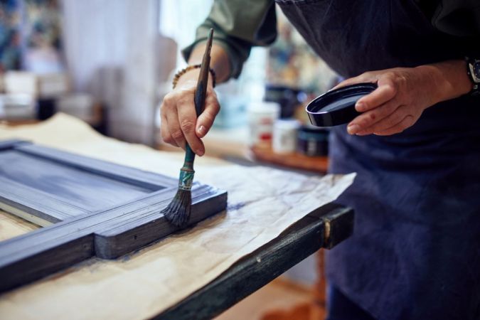 a person holding a brush and painting on a piece of wood in front of a window