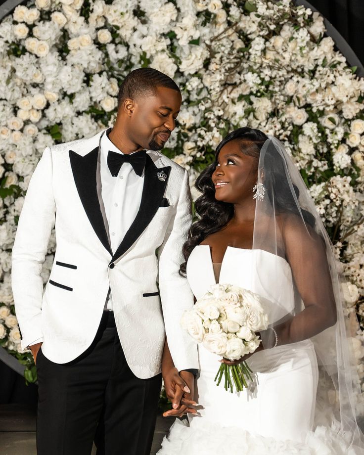 a bride and groom standing next to each other in front of a floral wall with white flowers