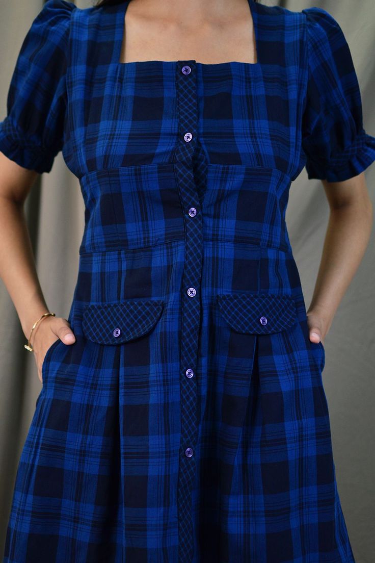 a woman is wearing a blue and black plaid dress with buttons on the front, she has her hands in her pockets