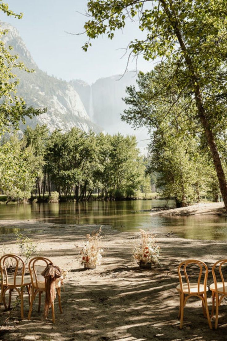 chairs and tables set up on the shore of a river with mountains in the background