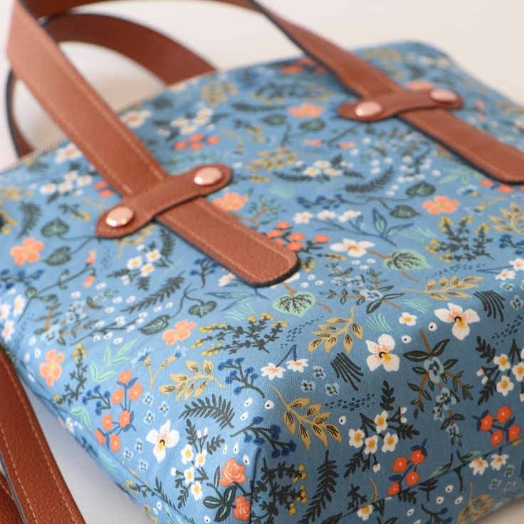 a blue flowered bag with brown straps on a white table and a tan leather strap