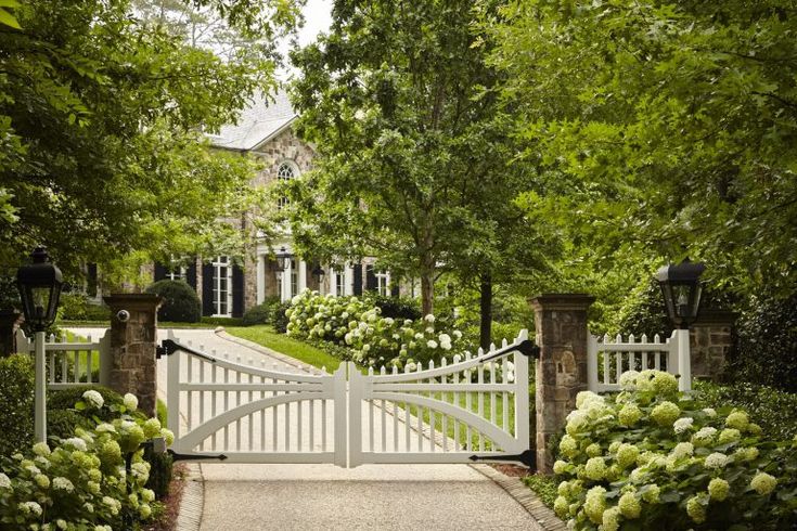 a white gate is surrounded by lush green trees and shrubbery in front of a large house