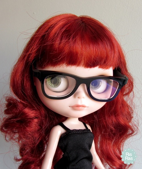 a doll with red hair, glasses and a black dress is standing in front of a white wall