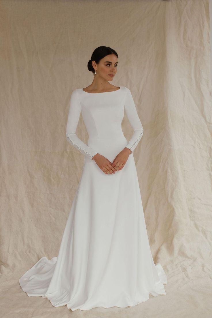 This unique wedding dress, made from luxurious crepe, is an ideal minimalistic elegance. With long sleeves and an A-line silhouette, this floor-length gown with a long train is perfect for winter weddings, including Christmas or civil ceremonies.  The modest design features a flattering boat neckline and a high back, creating a timeless and graceful look. Bridal buttons flow down the back and extend to the end of the train, adding a touch of classic beauty, long sleeves are also complemented wit Timeless Wedding Dress Modest, A Line Long Sleeve Wedding Dress, Classy Simple Wedding Dress, December Wedding Dress, Long Sleeve Wedding Dress Winter, Christmas Wedding Dress, Bridal Buttons, Wedding Dress Winter, Winter Wedding Dresses