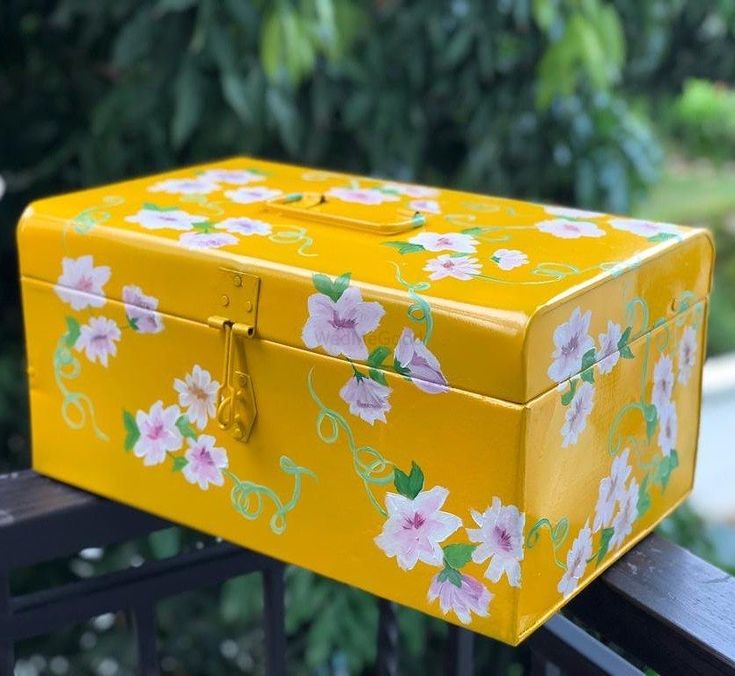 a yellow box with flowers painted on it sitting on a rail near some trees and bushes