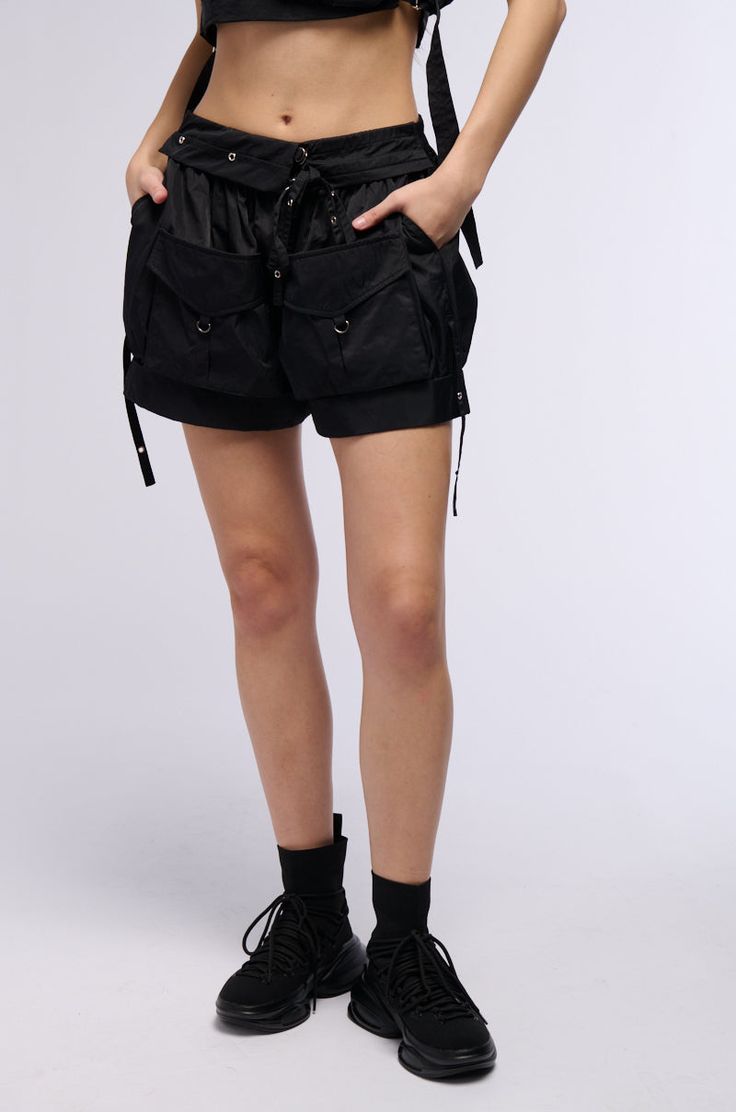 The AKIRA Label Kaden Cargo Mini Shorts In Black are made from a slick, non-stretch nylon fabrication and feature a high rise design with a shirred elastic waistband, fold over front-waist paneling, tonal stitching, faux silver and hematite metallic hardware, waist-pleat detailing, side slant front pockets, and dual front flap cargo pockets. Complete with grommet hardware, metallic D-ring accents, extended decorative side straps, a mini length silhouette with cuffed hems, a pull-on fit, and adjustable strappy buckle fastenings at the waist. Style with the coordinating AKIRA Label Kaden Cargo Sleeveless Top In Black to finish off your look.   - 100% Nylon - Non-Stretch Fabric; Stretchy Waistband - Imported  (all measurements are approximate from size small) - 12.5” Rise - 2.5” Inseam  - Mod Cargo Mini Shorts, Mini Shorts, Fold Over, Stretch Fabric, Sleeveless Top, High Rise, Black