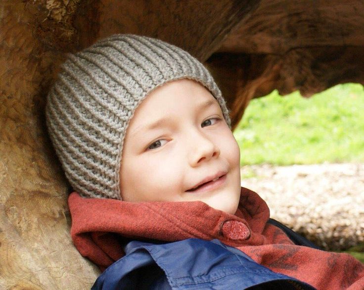 a young boy wearing a knitted hat and jacket under a large tree trunk in the shade