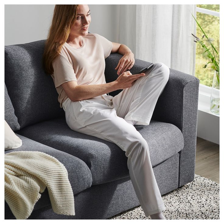 a woman sitting on top of a gray couch in front of a window with white curtains