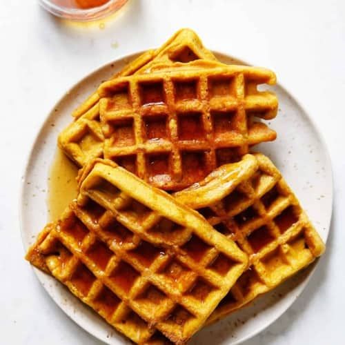 three waffles on a white plate with syrup and orange juice in the background