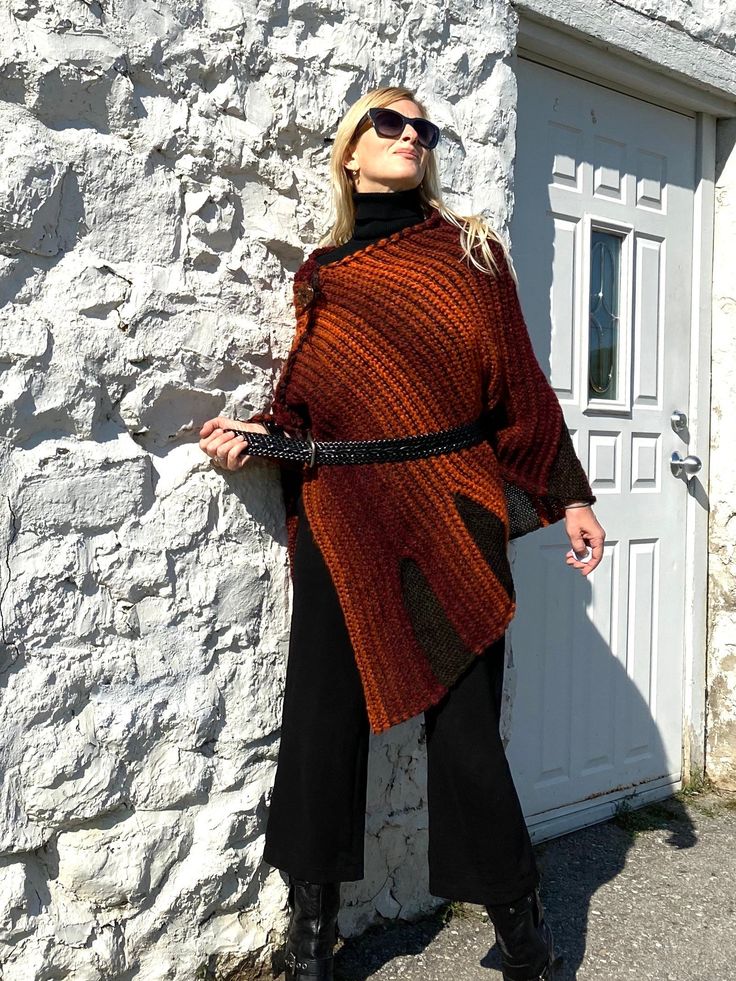 "Fall Knit Steampunk Ruffle Wrap | Boho Alpaca Black Orange Poncho, Wool Poncho Cape For Women This flirty, ruffled, alpaca wool blend hand-knitted wrap in black and burnt orange tons, is light and warm. It is a four-season piece and is ideal for layering.  The colour palette of this versatile shawl is definitely jeans friendly.  STYLE This dressy/casual piece is an excellent travel companion.  Works well with jeans, leggings, dress pants or over a solid dress adding a flowy elegance of a shawl. Endless in its versatility and fits most sizes make it a great gift idea. Material: Alpacas Wool Blend WAYS TO WEAR SEE FAQ's FOR WAYS TO WEAR AS A SHAWL, FITTED LOOK, BELTED LOOK AND SCARF. SIZING Length is approx. 30\" Fits sizes S- 3xl Please inquire about larger sizes Our model Elana is size 8 Acrylic Poncho Shawl For Fall, Acrylic Shawl Poncho For Fall, One Size Acrylic Poncho For Fall, One Size Cape For Fall Festival, Bohemian Black Poncho For Fall, Fall Festival Cape Poncho, Hand Knitted Shawl Poncho For Fall, Bohemian Knitted Poncho For Fall, Knitted Shawl Cape For Fall