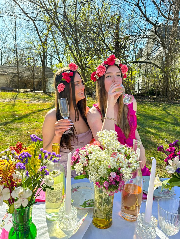 two women sitting at a table with flowers in their hair drinking wine and holding glasses