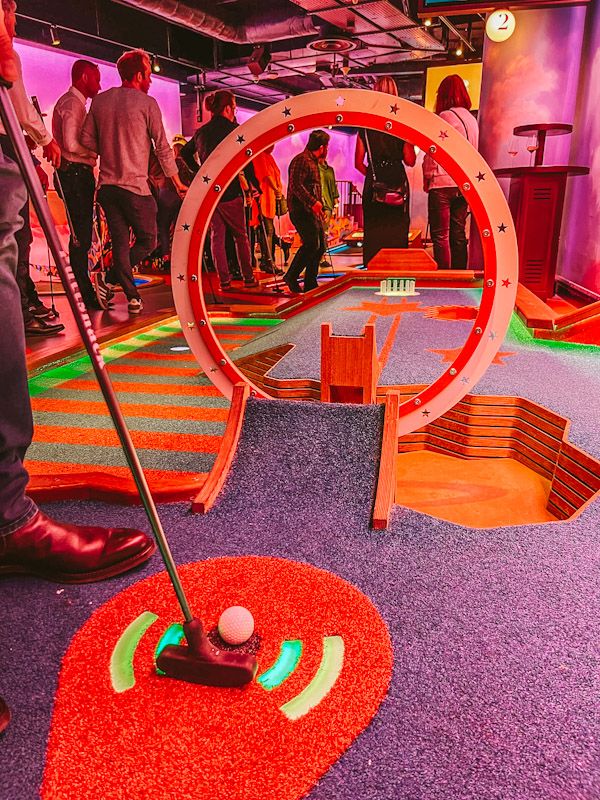 people are standing around playing miniature golf in an indoor area with red carpet and colorful lights