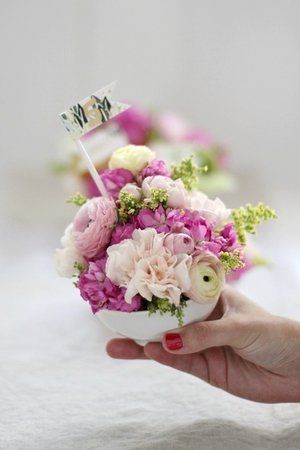 a hand holding a white bowl filled with pink and white flowers on top of a table