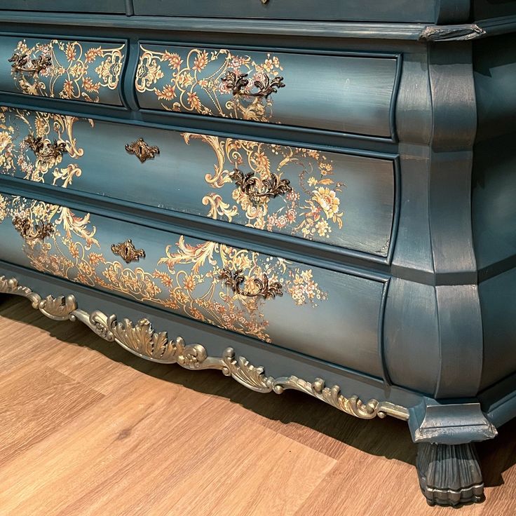 an ornate blue dresser with gold paint on it's drawers and drawer pulls, sitting on a wooden floor