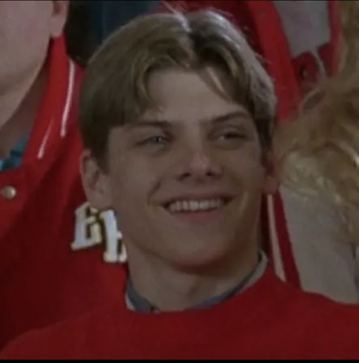 a young man wearing a red uniform and smiling at the camera with other people behind him