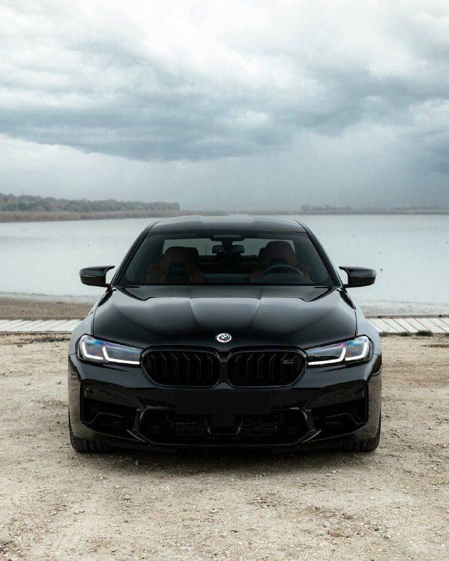 the front end of a black bmw car parked on dirt near water and cloudy sky