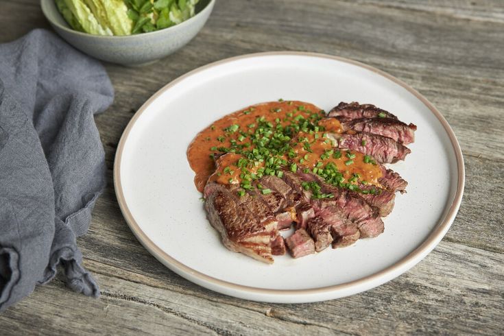 a white plate topped with meat covered in sauce next to a bowl of lettuce