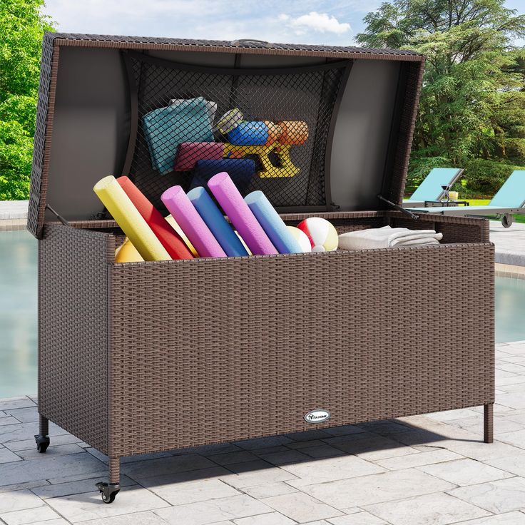 an outdoor wicker storage box with pool toys in the lid and on wheels, sitting next to a swimming pool