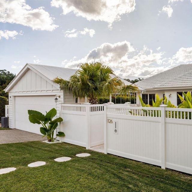 a white fence in front of a house with palm trees and bushes on the lawn