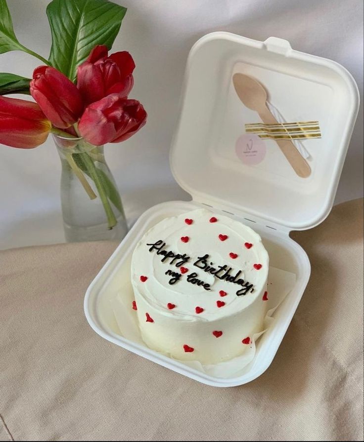 a white cake in a plastic container next to a vase with red tulips