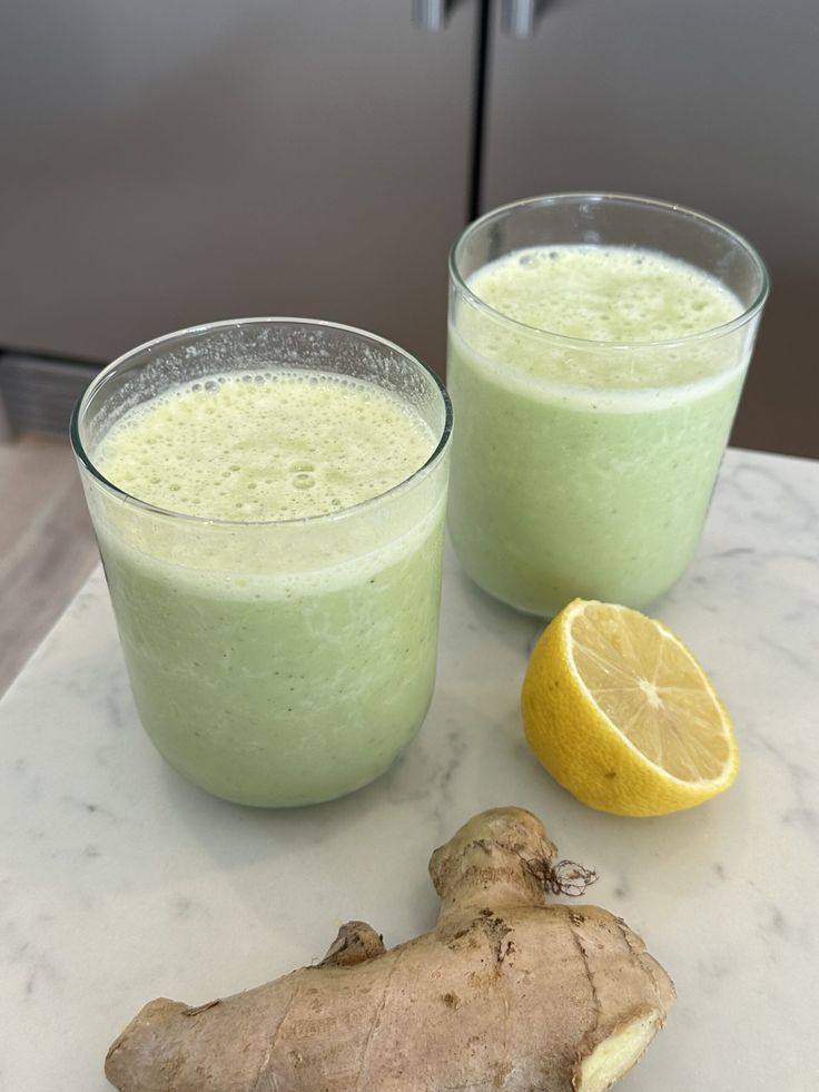 two glasses filled with green smoothie next to a sliced lemon on a marble counter