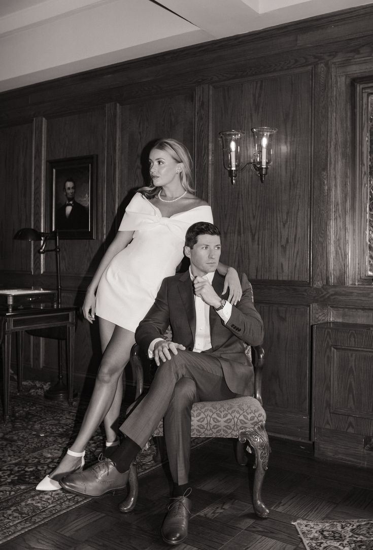 black and white photograph of man sitting on chair with woman standing next to him in formal dress