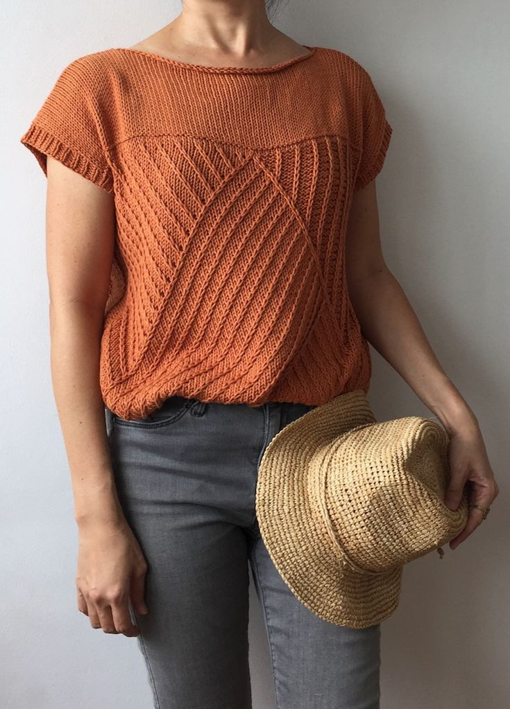 a woman standing next to a wall with a hat in her hand and wearing an orange sweater