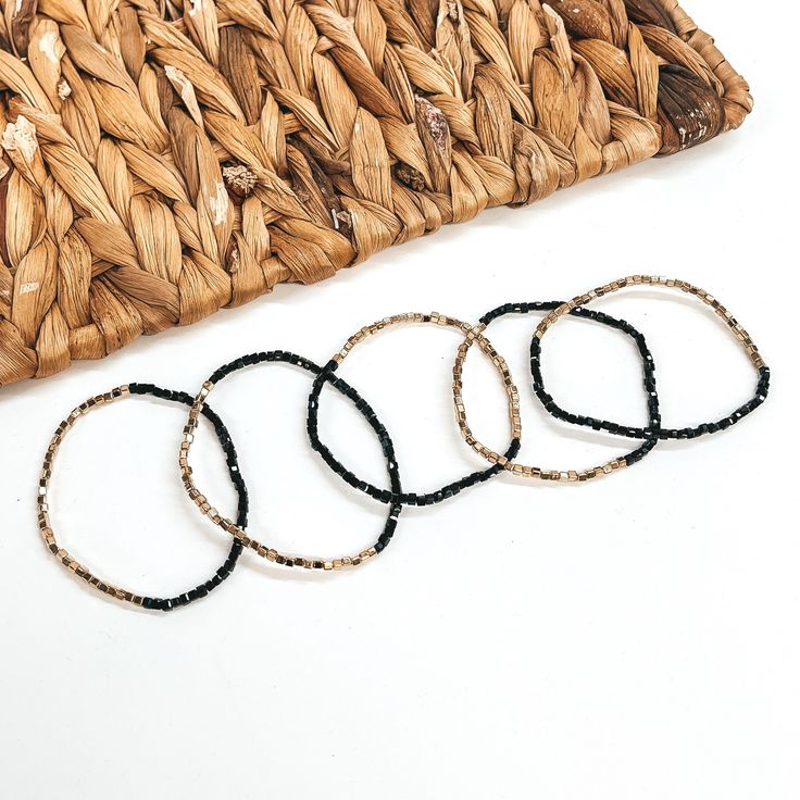 These are five seed beaded bracelets with gold and  black shiny beads. They are taken on a white  background and a brown woven plate in the back as  decor. Trendy Black Friendship Bracelets For Beach, Trendy Adjustable Beaded Bracelets With Black Beads, Trendy Black Bracelets For The Beach, Trendy Black Bracelets For Beach, Minimalist Beaded Bracelets With Spacer Beads For Beach, Trendy Adjustable Bracelet With Black Beads, Minimalist Beaded Bracelets For Beach With Spacer Beads, Trendy Black Beaded Bracelets With Tiny Beads, Minimalist Beaded Bracelets With Spacer Beads