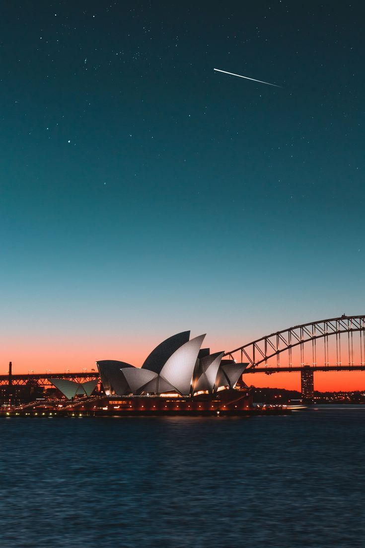 the sydney opera house is lit up in front of an illuminated bridge at night with a shooting star above it