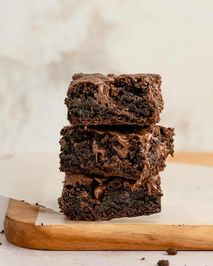 three chocolate brownies stacked on top of each other next to a wooden cutting board