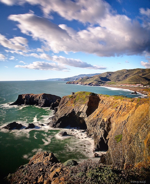 an ocean view with waves crashing on the shore and cliffs in the foreground, under a cloudy blue sky