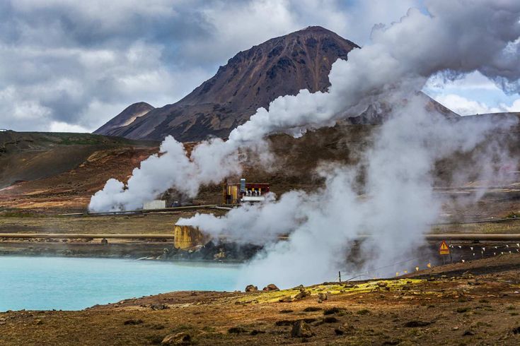 steam billowing out from the ground near a body of water with mountains in the background