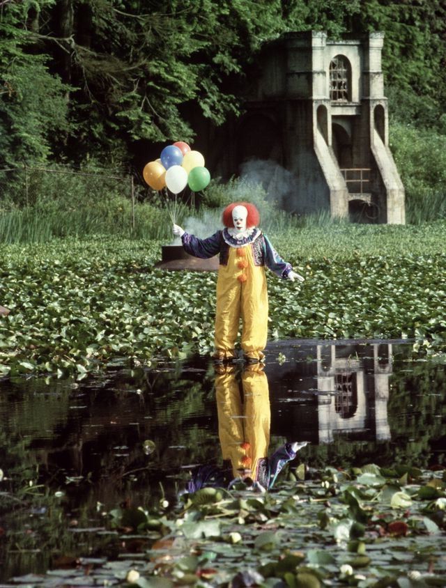 a clown in yellow jumpsuit holding balloons near water with lily pads on the ground