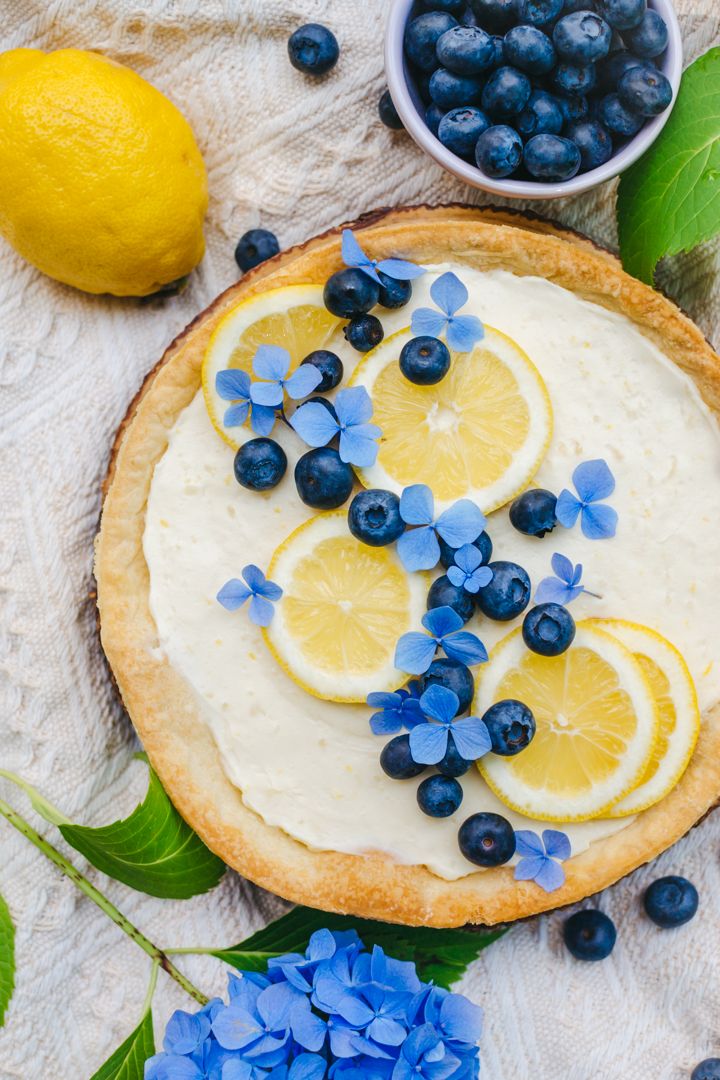 a pie with lemons and blueberries on it next to some flowers, leaves and water