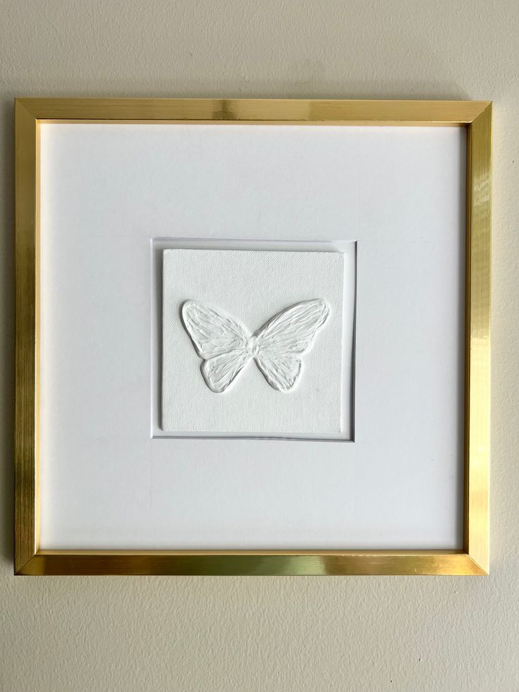 a white and gold framed artwork with a butterfly on it's back, hanging on the wall