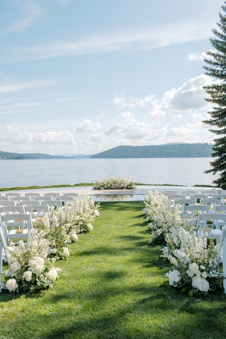 an outdoor ceremony set up with white chairs and flowers on the grass by the water