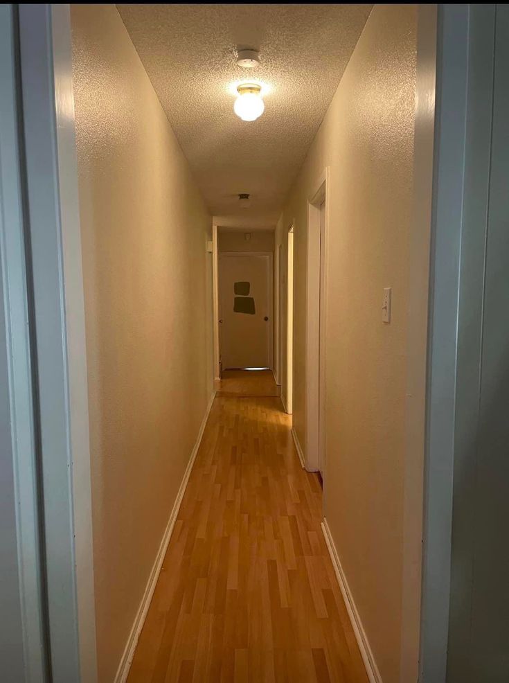 an empty hallway with hard wood flooring and light fixture on the far wall, leading to another room