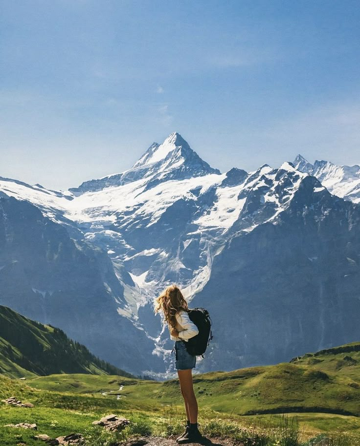 a woman standing on top of a lush green hillside next to a snow covered mountain