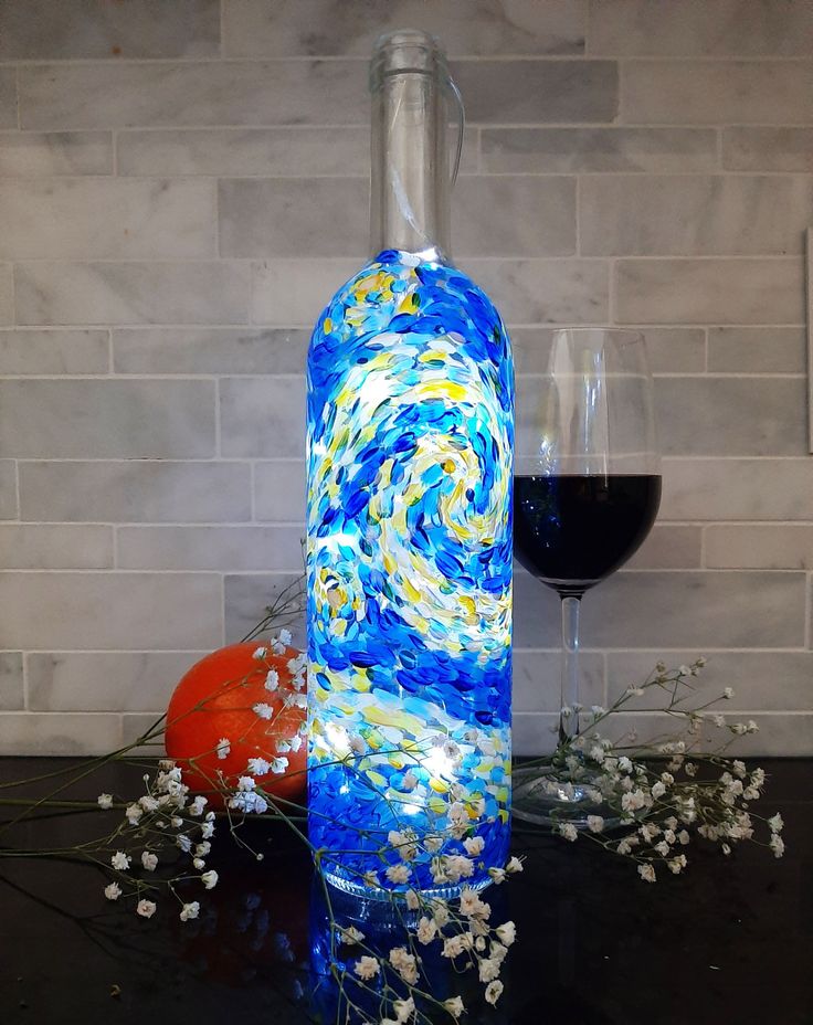 a wine bottle that has been painted with blue and white swirls, next to a glass of red wine