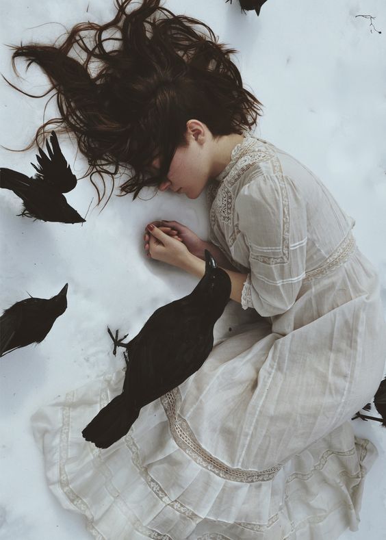 a woman is sitting in the snow with her head down and birds flying around her