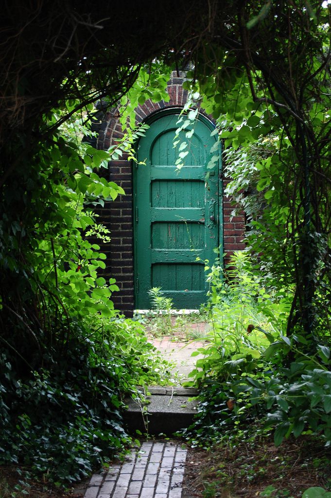 an open green door surrounded by greenery and brick walkway leading to the entrance way