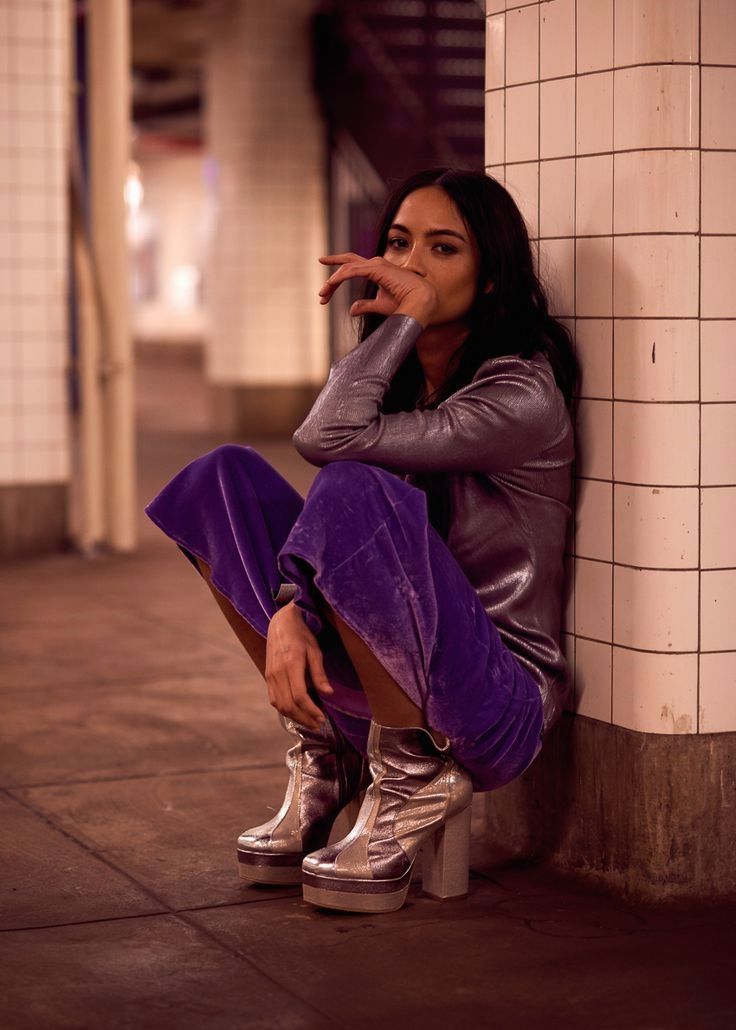 a woman sitting on the side of a building next to a white brick wall with her hand under her chin