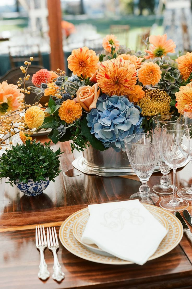 an arrangement of flowers and greenery is displayed on a wooden table with silverware