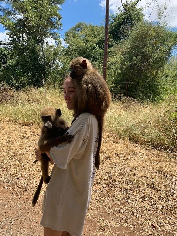 a woman holding a monkey on her back in the middle of a dirt road with trees and bushes behind her