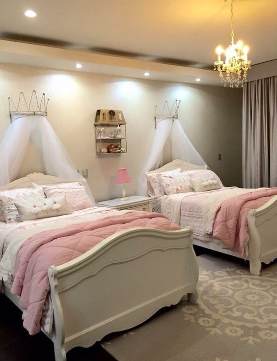 two white beds in a room with pink comforters and pillows on them, one has a chandelier hanging from the ceiling