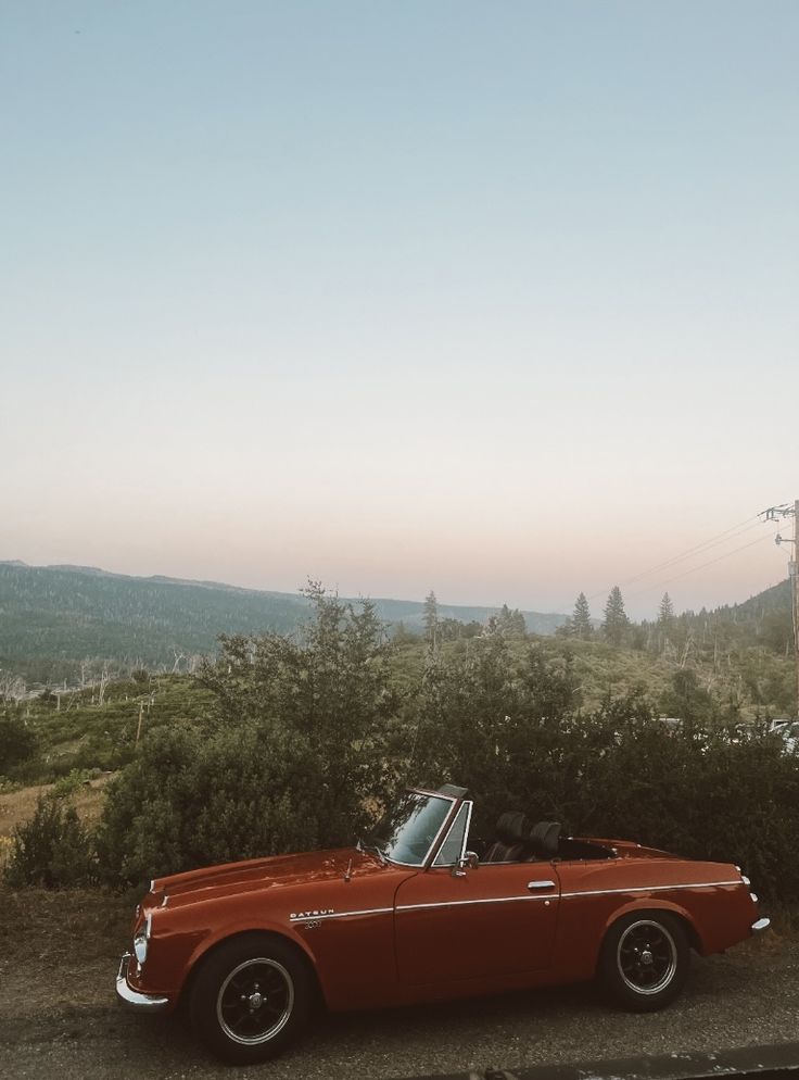 an old red convertible car parked on the side of the road in front of some trees